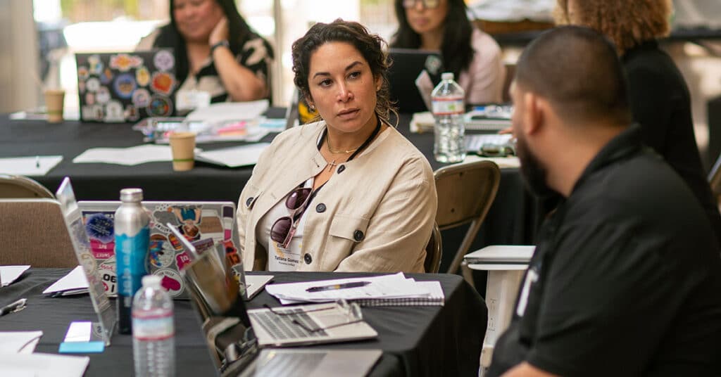 A woman sits at a conference table covered with laptops, papers, and water bottles, attentively listening to senior leaders speaking. Other people work and talk in the background.