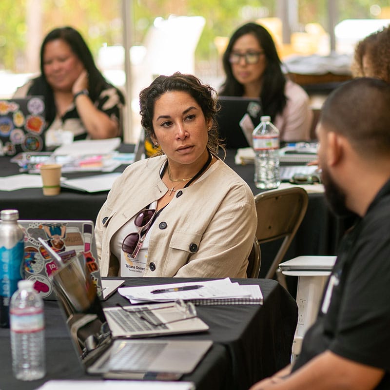 A woman with curly hair, wearing a light jacket and sunglasses around her neck, sits at a table with laptops and notebooks, listening intently to senior leaders. Others work in the bright room, focused on improvement reviews.