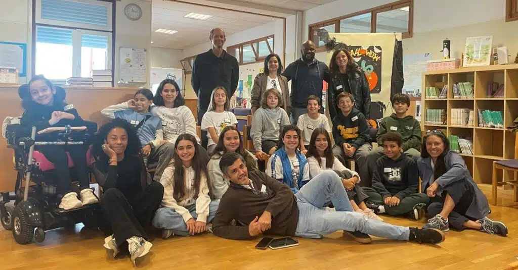 A group of students and adults pose together in a classroom in Oeiras, some sitting on the floor and others standing behind. Bookshelves and posters are visible in the background, reflecting a learner-centered education. Everyone appears relaxed and happy.