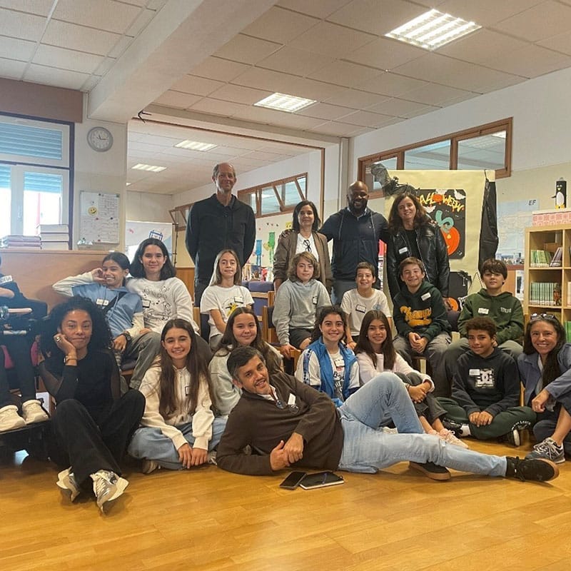 A group of adults and children pose together and smile in a bright Oeiras classroom filled with bookshelves and posters. Some are sitting on the floor, while others are seated or standing behind them, reflecting a learner-centered education environment.