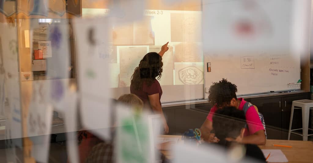 A teacher points at a projection on a classroom whiteboard while students engage in deeper learning at their desks; the scene is viewed through a glass wall decorated with paper drawings.