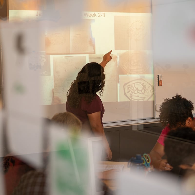 A teacher stands at the front of a classroom, pointing to images and text projected on a whiteboard, engaging students in deeper learning as they watch attentively; reflections on glass partially obscure the scene.