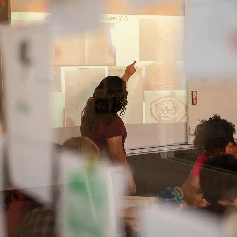 A teacher stands at the front of a classroom, pointing to images and text projected on a whiteboard, engaging students in deeper learning as they watch attentively; reflections on glass partially obscure the scene.
