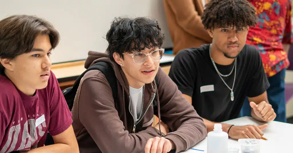 Three young men sit at a table, attentively listening. Wearing casual clothes and name tags, they have notebooks and bottles in front of them. The classroom setting suggests a focus on class participation among multilingual learners.