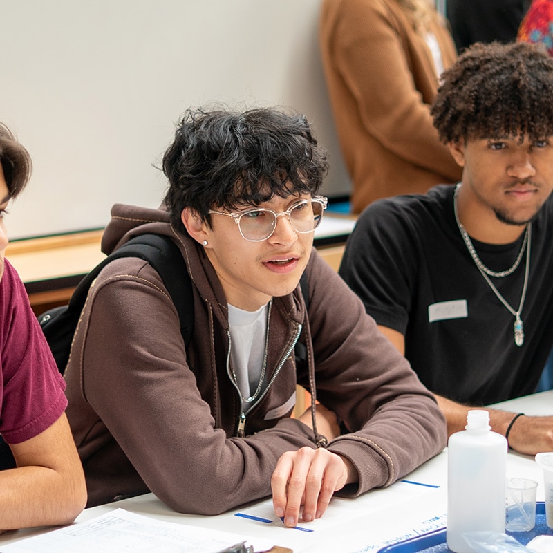 Two young men sit at a table with papers and a water bottle, listening attentively. Their engaged expressions suggest active class participation in a workshop, possibly focused on Improvement Science or supporting multilingual learners.