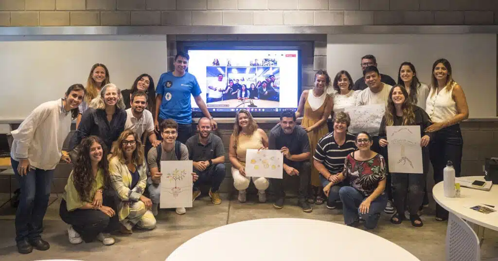 A group of people pose and smile in a classroom, some kneeling, some standing. Three hold up collaborative drawings—highlighting their Professional Learning Community. Behind them, a large screen shows a video call with more members. The setting appears friendly and informal.