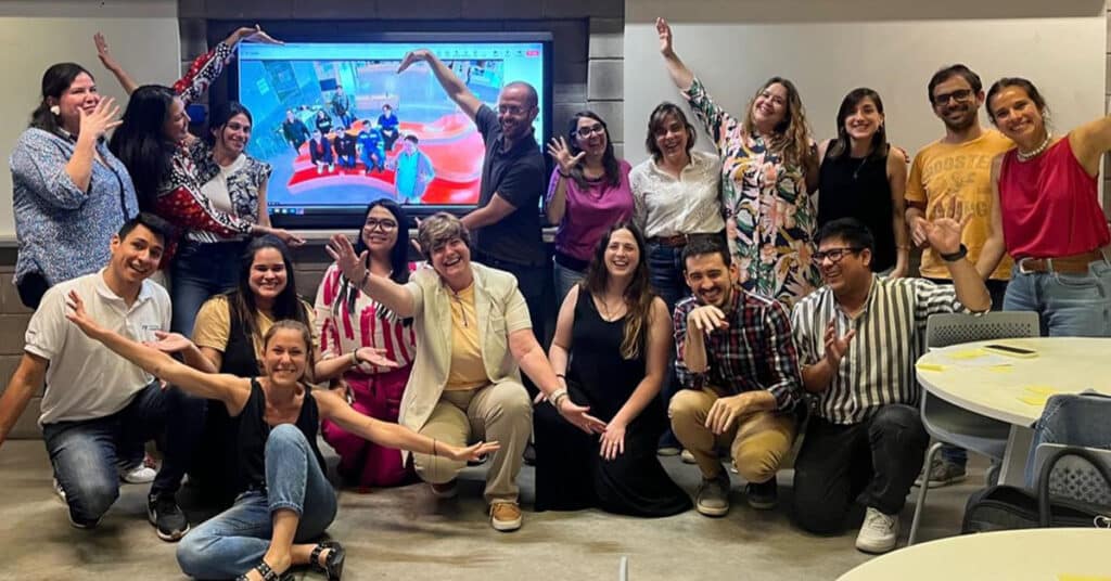 A group of people pose cheerfully in a classroom, showcasing how Professional Learning Communities can transform teaching. Some sit and others stand before a colorful screen, all smiling and making playful gestures with their arms.