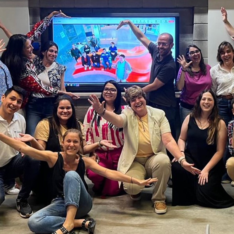 A group of people pose cheerfully in front of a screen displaying a video call with more smiling participants, some raising their arms or making heart shapes, as they engage in professional learning communities in a bright indoor setting.