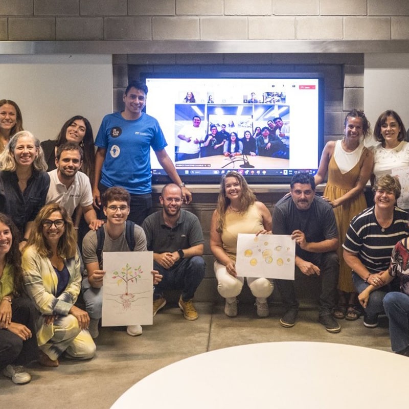 A group of people smiling and posing indoors in front of a screen with a video call, embodying Professional Learning Communities. Some in the front row hold hand-drawn posters. The casual setting fosters collaboration and shared growth.