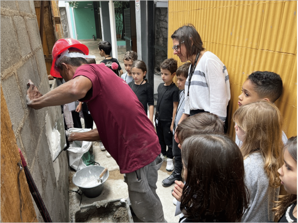 A group of first graders and an adult watch a construction worker in a red hard hat apply plaster to a cinder block wall at a building site, part of a co-designed project with the community. A yellow wall and construction materials are visible nearby.