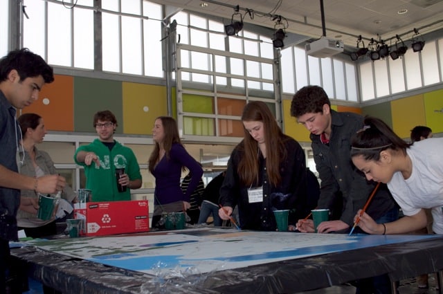A group of young people work together on the Imagine Mural project in a bright, modern room. Some are painting on a canvas to honor a life, while others watch and talk. Paint cups and supplies are spread across a table covered with plastic.
