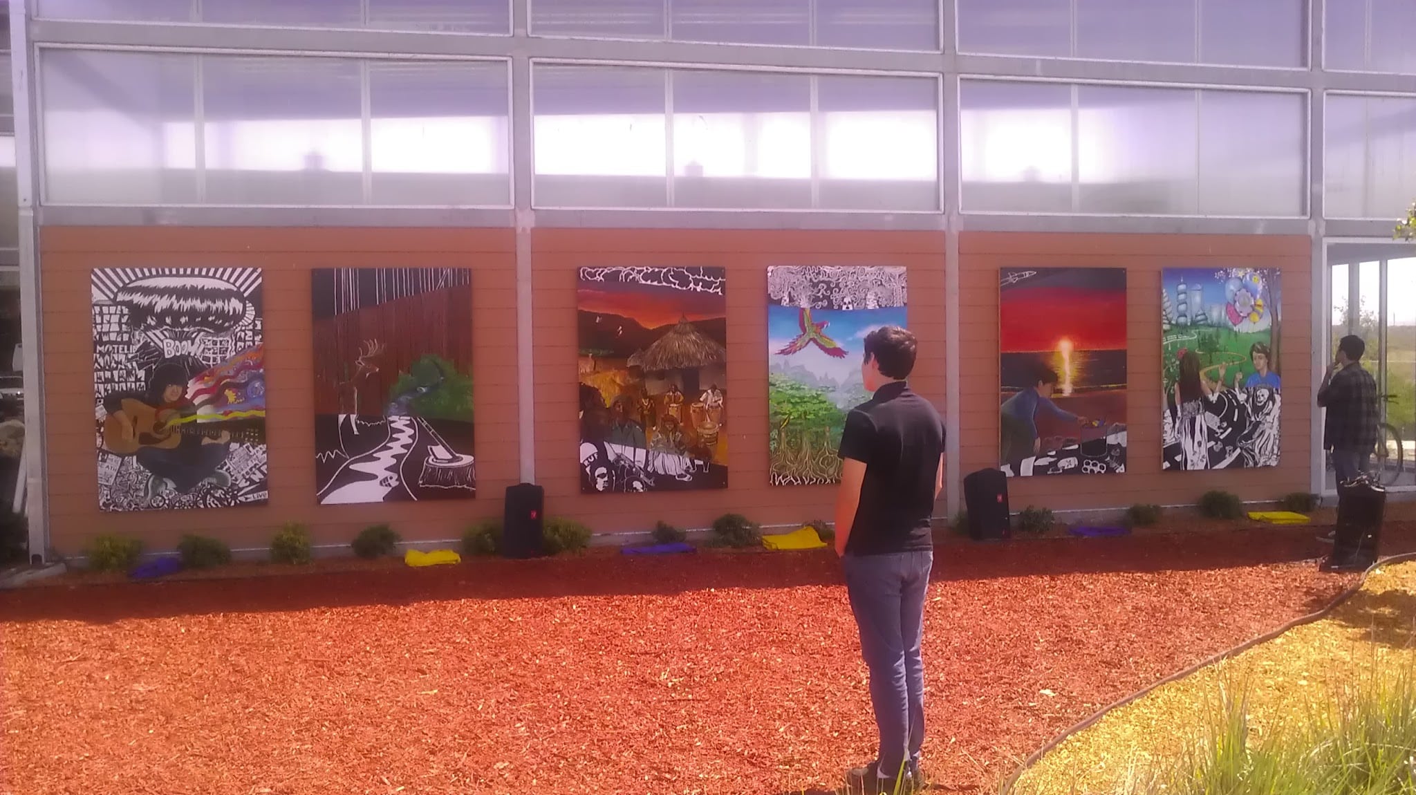 A person stands in front of a wall displaying five colorful paintings outdoors, part of the Imagine Mural project. Another person appears near the edge, with mulch on the ground and plants by the wall honoring a life through art.
