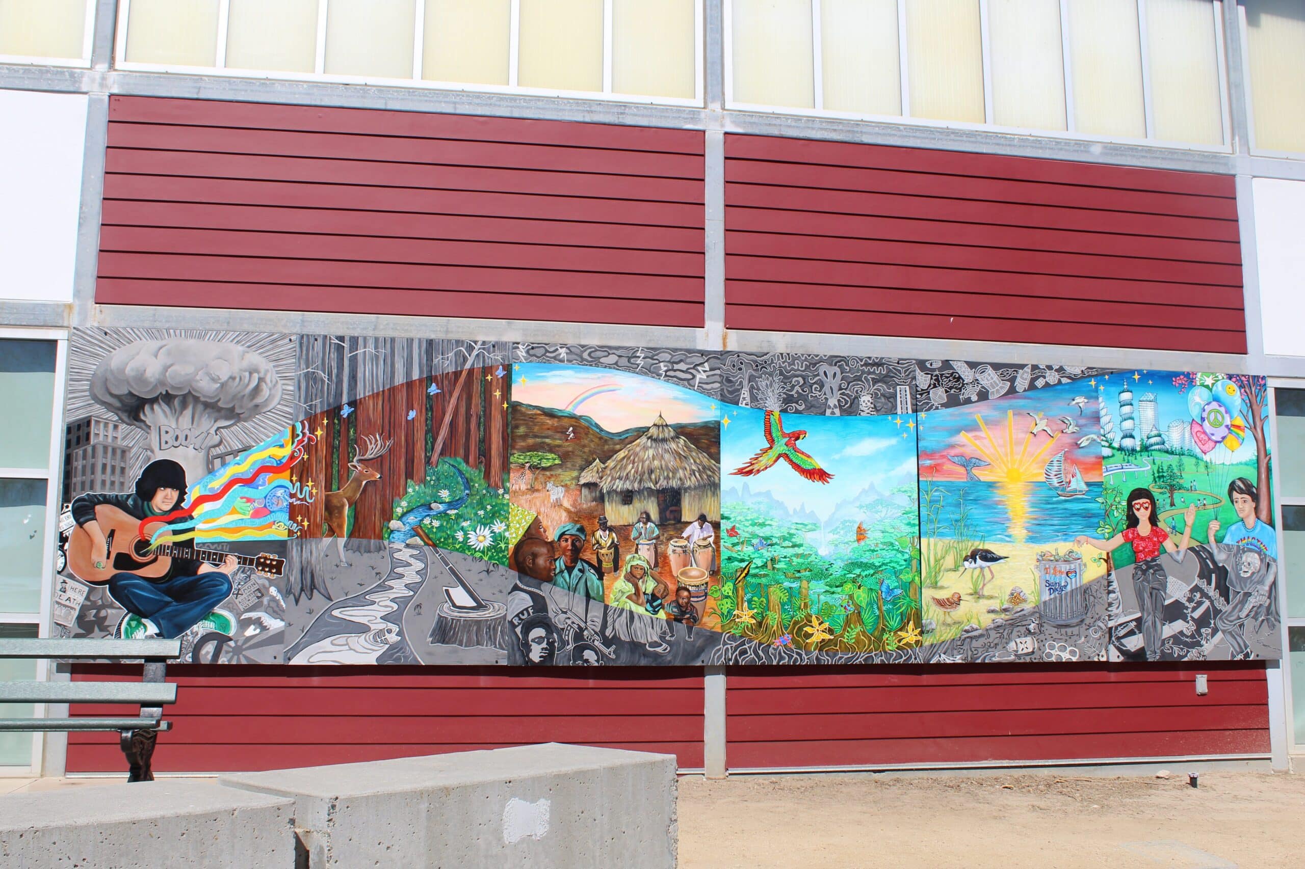 A colorful Imagine Mural on a red and white building depicts a person playing guitar, cultural and natural scenes, animals, and vibrant landscapes, blending grayscale and bright colors to honor a life through music, nature, and community themes.
