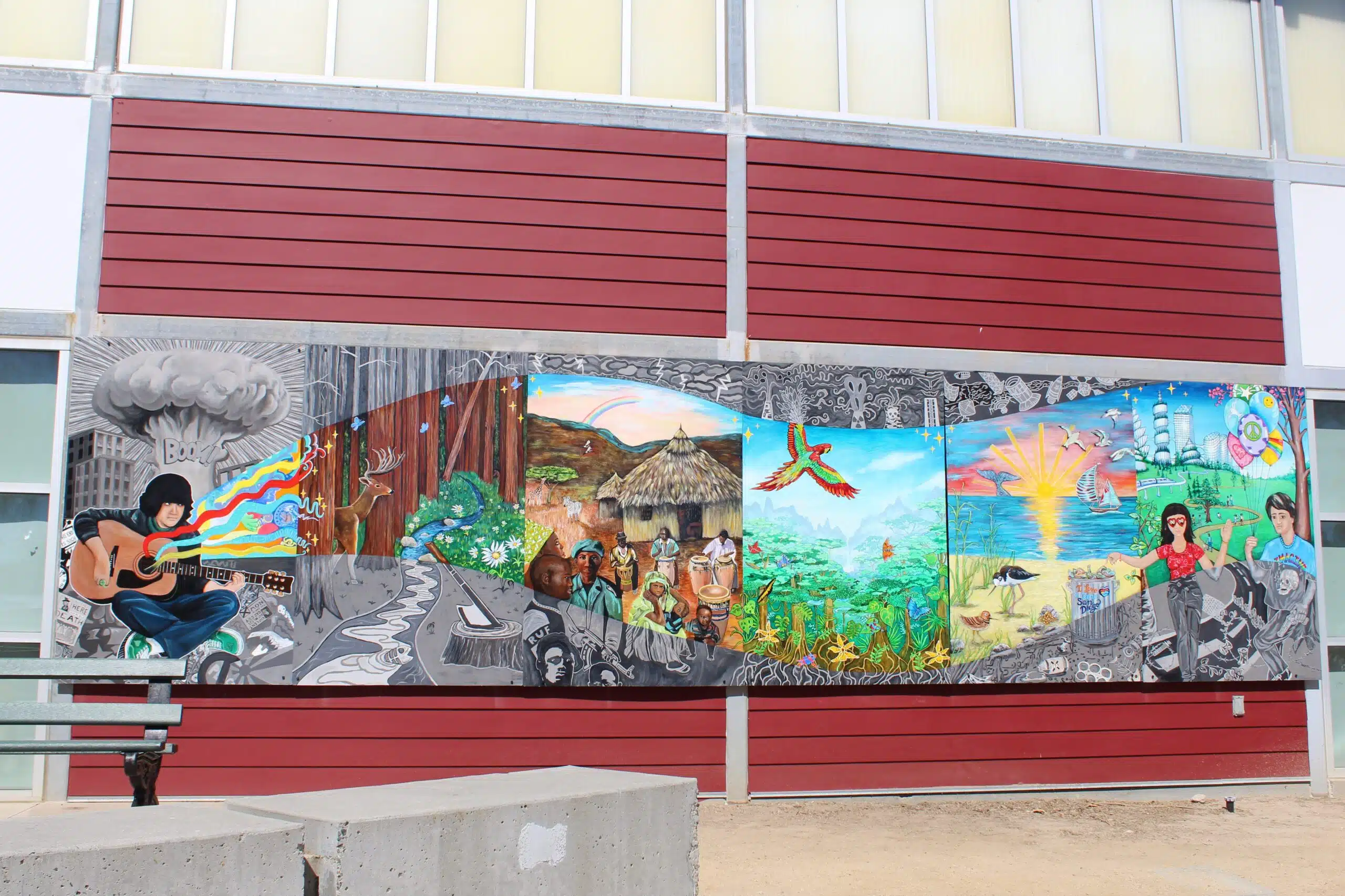 A colorful Imagine Mural on a red and white building depicts a person playing guitar, cultural and natural scenes, animals, and vibrant landscapes, blending grayscale and bright colors to honor a life through music, nature, and community themes.
