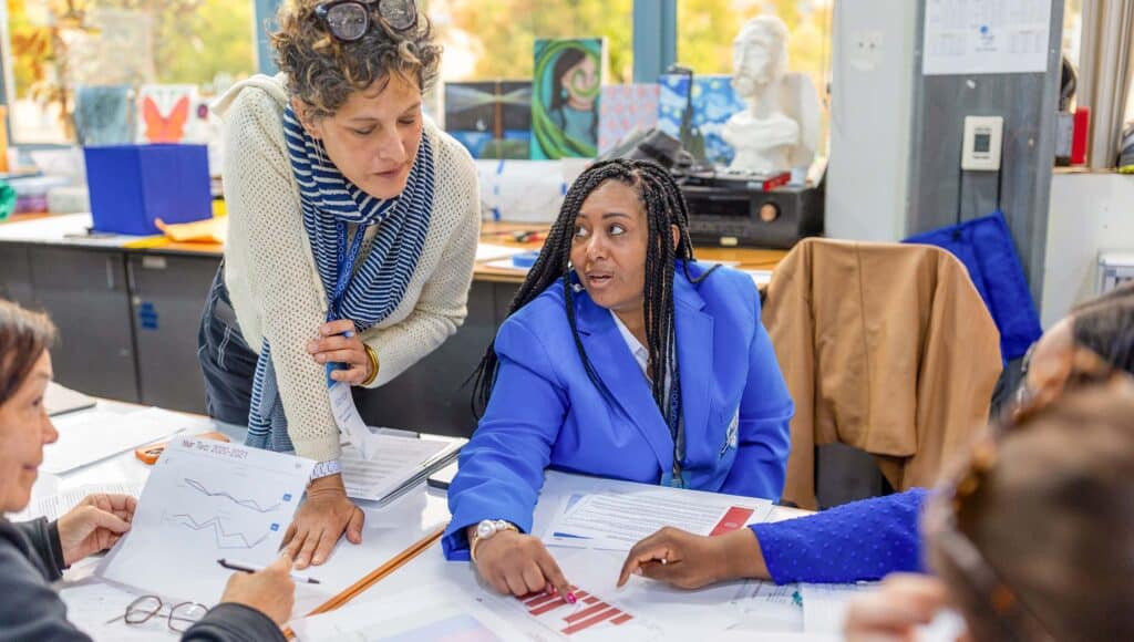 Two women, one standing and one seated, discuss documents and charts with graphs at a table during a meeting. Other people are partially visible, and office supplies are in the background.