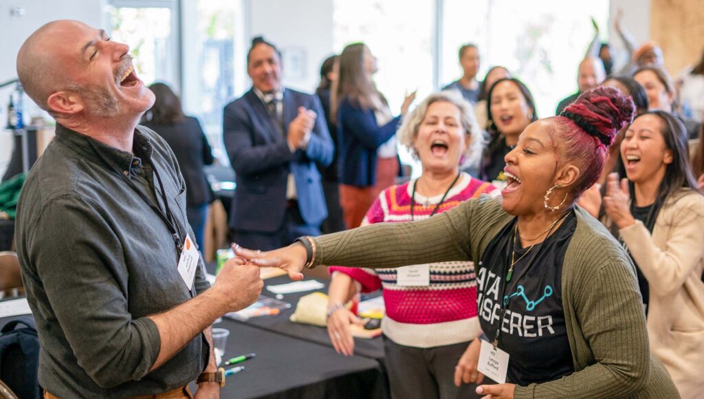 Two people in the foreground laugh and interact energetically in a lively, diverse group setting, while others in the background smile, clap, and cheer, suggesting a joyful, engaging event or workshop.