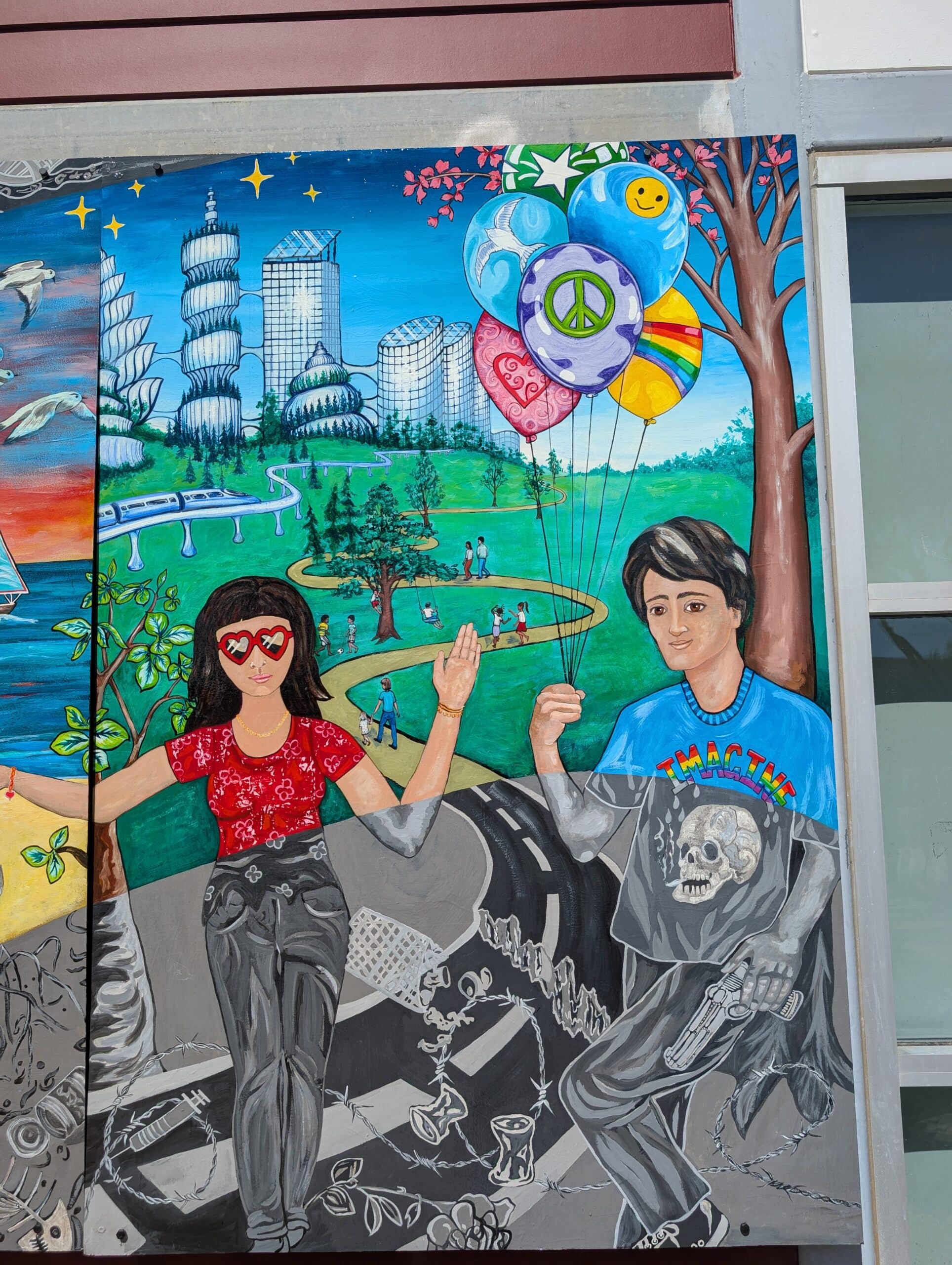 A colorful Imagine Mural depicts a girl wearing heart-shaped sunglasses and a boy in a graphic T-shirt holding balloons with peace symbols, set in a vibrant park with futuristic buildings and a grayscale lower section—a true Project to Honor A Life.