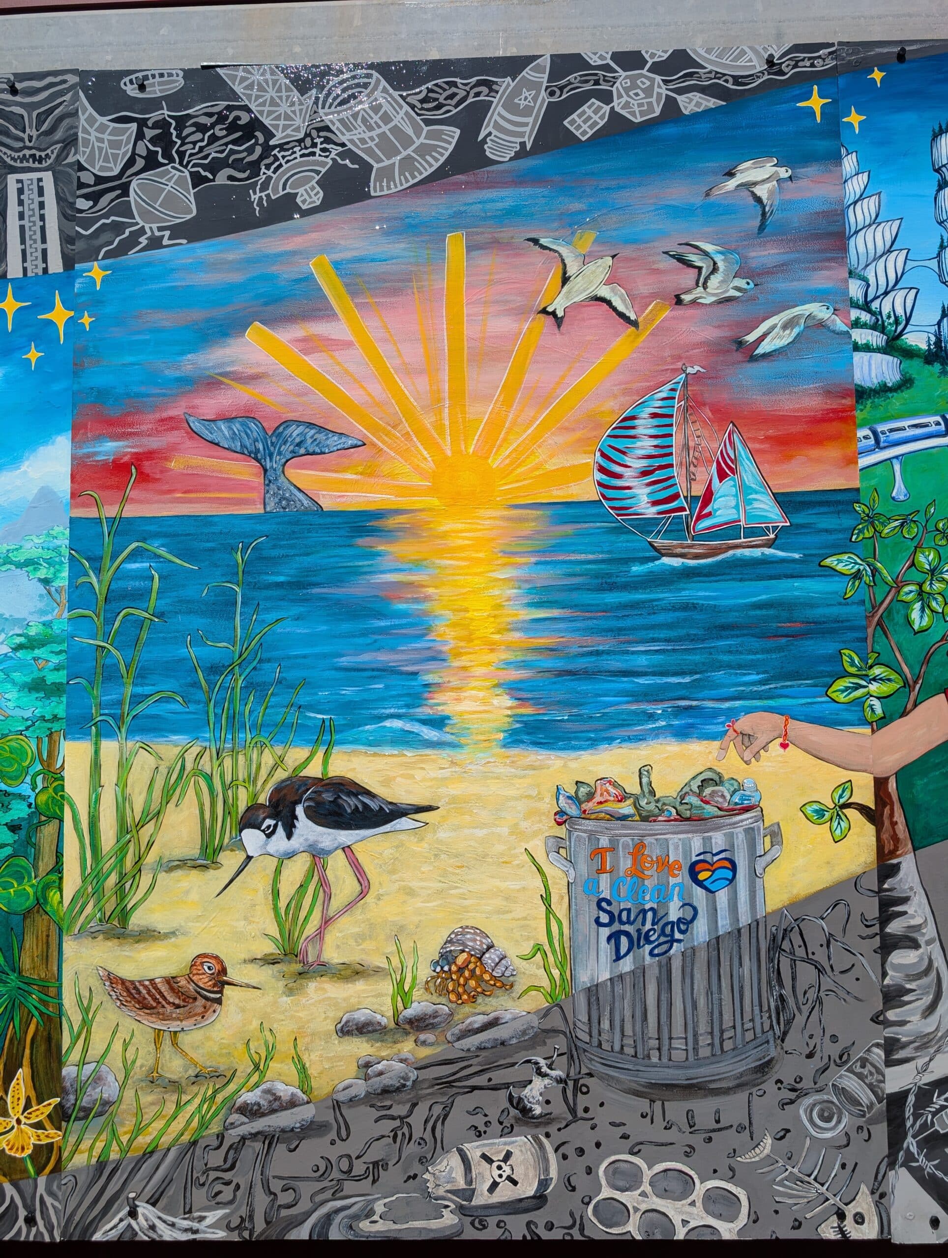A colorful Imagine Mural shows a sunset over the ocean with a whale tail, birds, and a sailboat. In the foreground, shorebirds walk on sand near a trash can labeled I Love Clean San Diego filled with litter.
