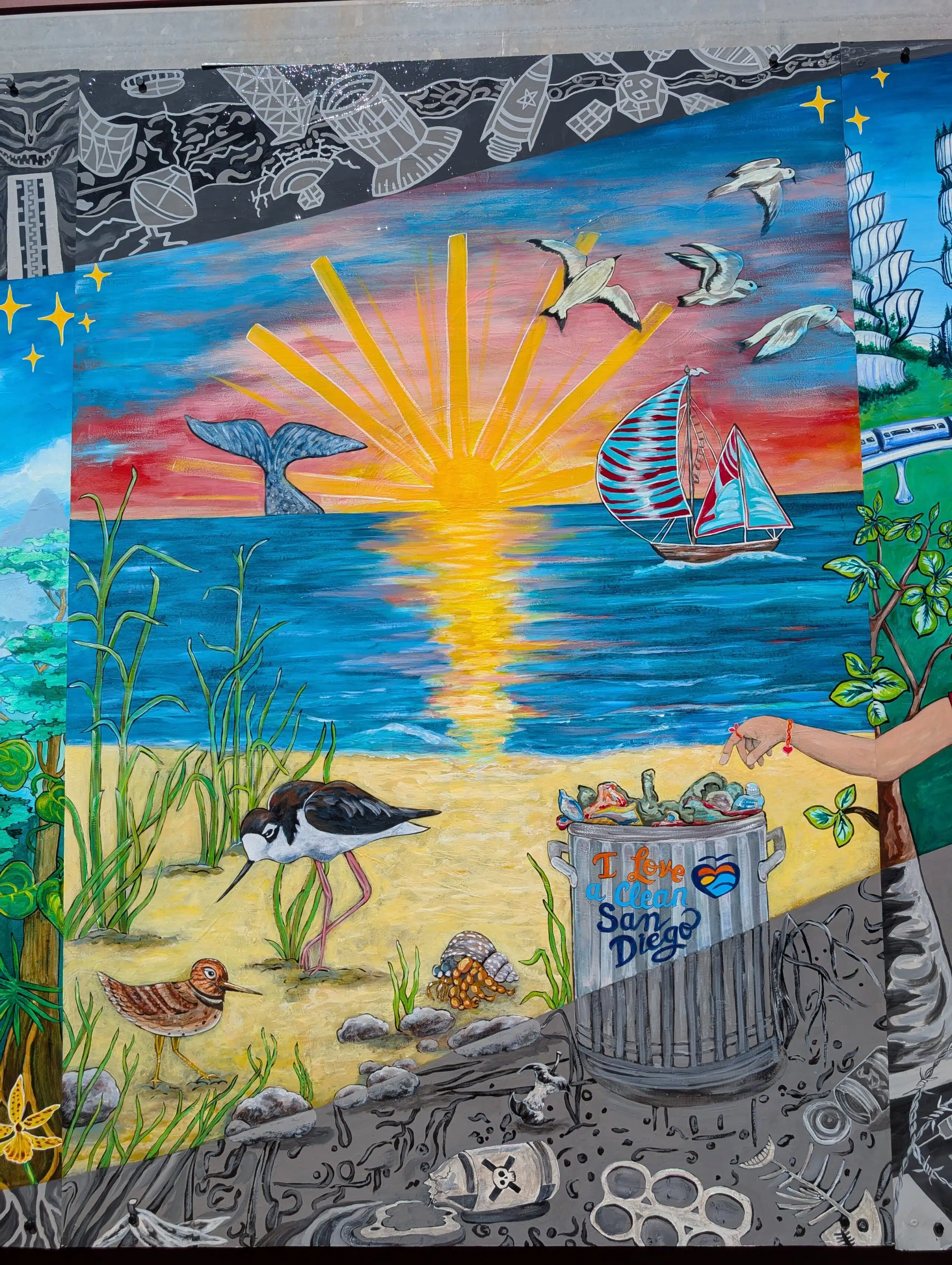 A colorful Imagine Mural shows a sunset over the ocean with a whale tail, birds, and a sailboat. In the foreground, shorebirds walk on sand near a trash can labeled I Love Clean San Diego filled with litter.