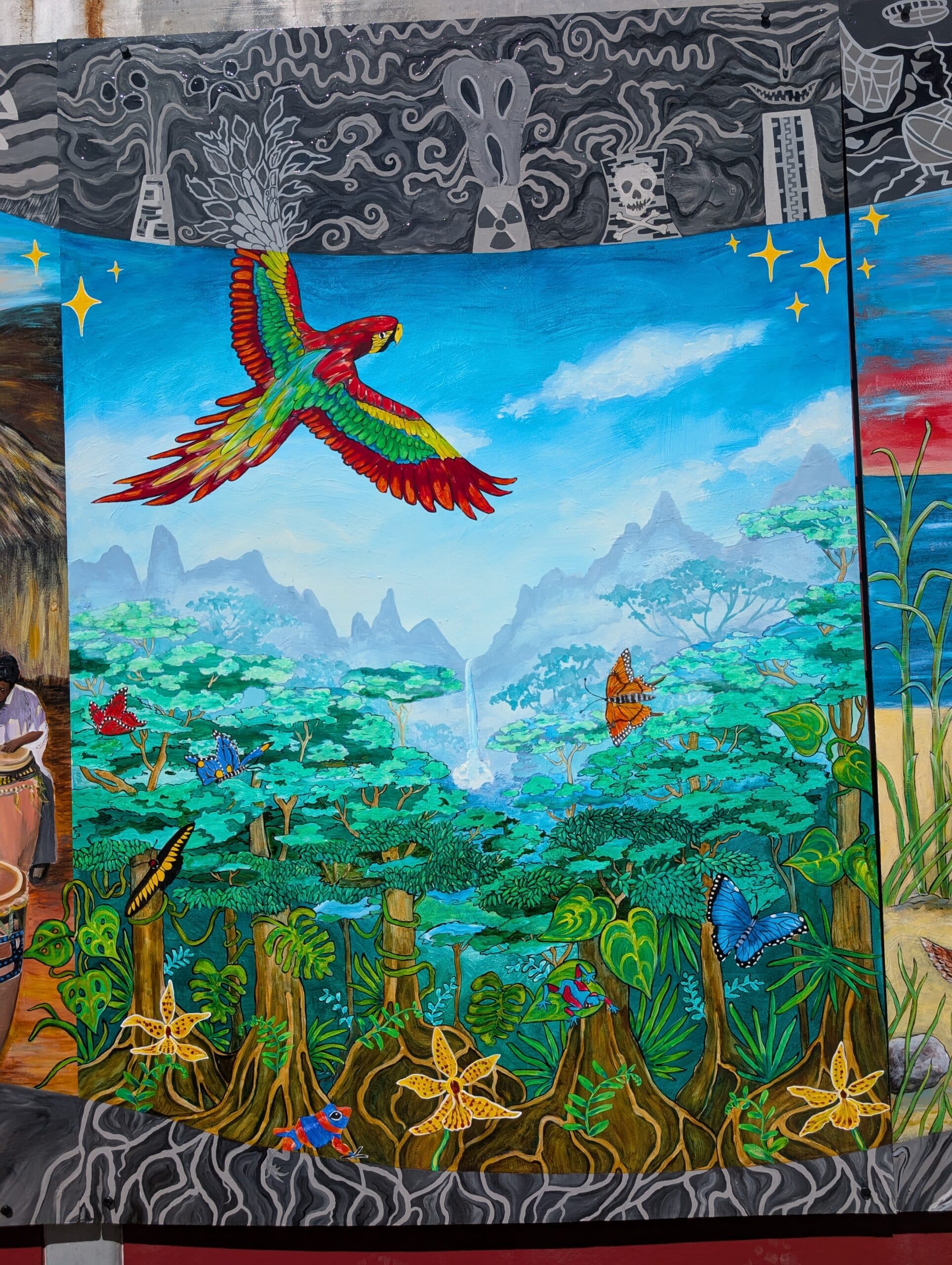A vibrant Imagine Mural depicts a red and green parrot flying above a lush tropical rainforest with butterflies, colorful flowers, green trees, and distant mountains under a bright blue sky, created to honor a life through art.