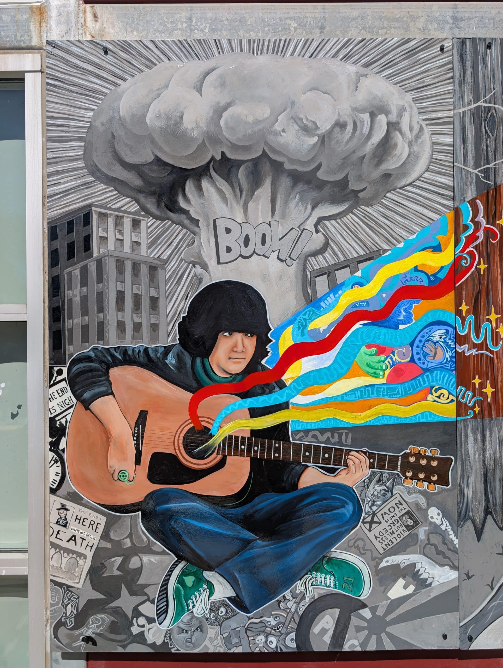 A mural from the Imagine Mural project shows a person with black hair playing guitar as colorful soundwaves burst out. A mushroom cloud and the word BOOM! rise behind, while city buildings and chaotic imagery fill the grayscale background.