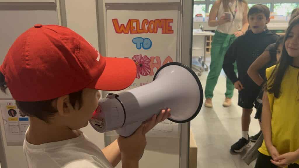A first grader in a red cap speaks into a megaphone in front of a colorful Welcome to sign, while other children and an adult listen, showcasing their co-designed project in a vibrant classroom community.