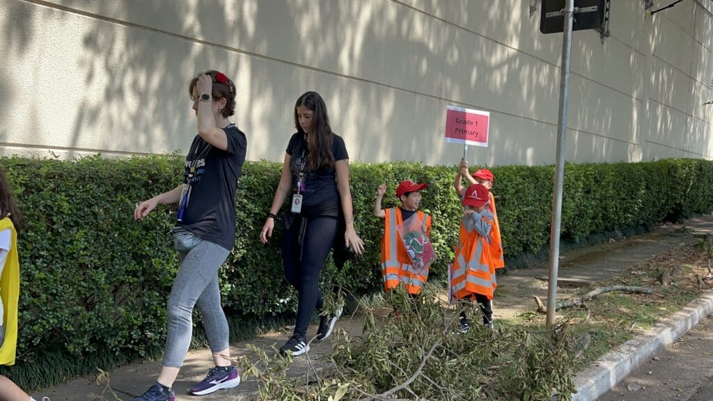 A group of first graders in orange reflective vests and red hats walk on a sidewalk with two adults. One child holds a pink sign that reads Grade 1 Pharmacy. This co-designed project brightens the path lined with shrubs and tree branches.