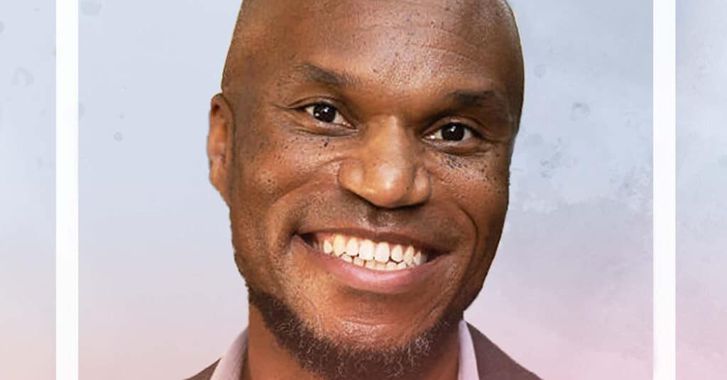 Cornelius Minor, a smiling man with a bald head and a closely trimmed beard, wearing a suit jacket and shirt, poses against a soft, light-colored background.