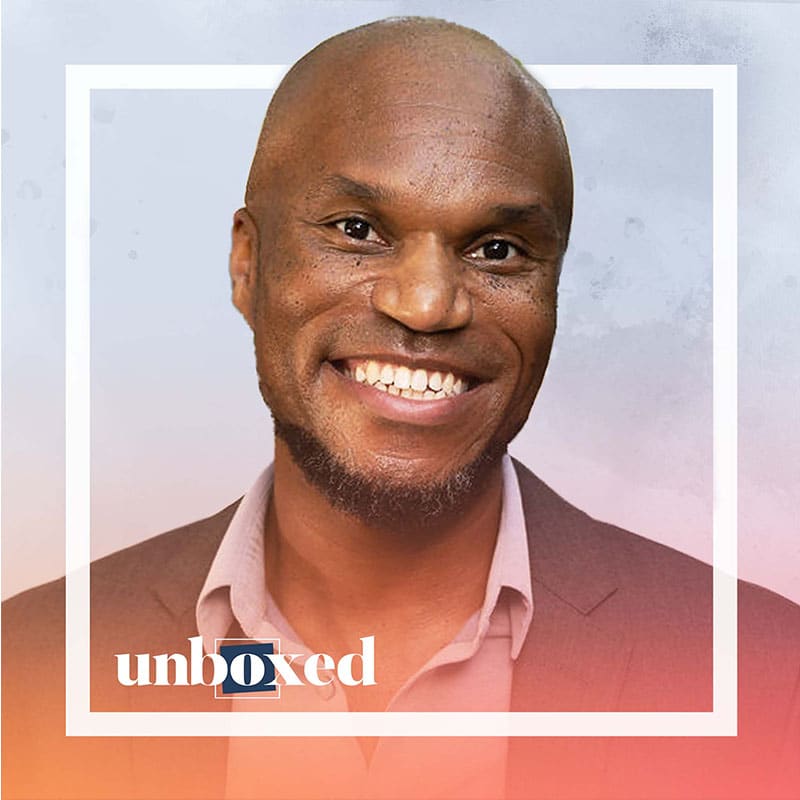 A smiling Cornelius Minor in a suit jacket and open-collared shirt is framed by a white square with the word unboxed at the bottom, on a soft gradient background.