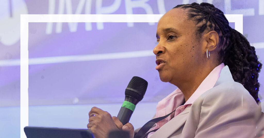 A woman in a light-colored blazer speaks into a microphone during the Mississippi Miracle event. She has braided hair tied back and is shown in profile against a purple and white background.