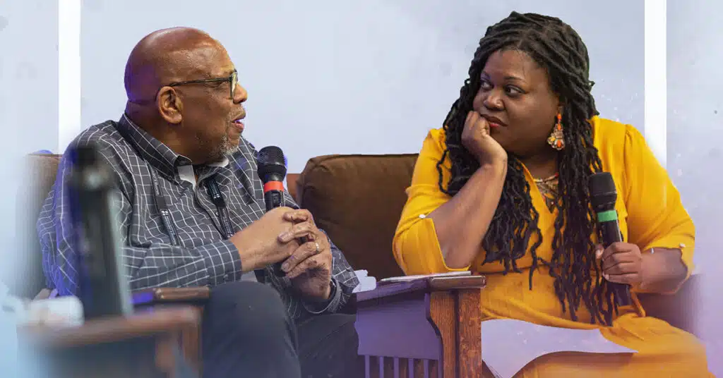 Two people sit and talk on stage, each holding a microphone. The man on the left wears glasses and a plaid shirt; Brandi Hinnant-Crawford, in a yellow dress, listens intently with her hand on her chin, guiding resistance and promoting change.