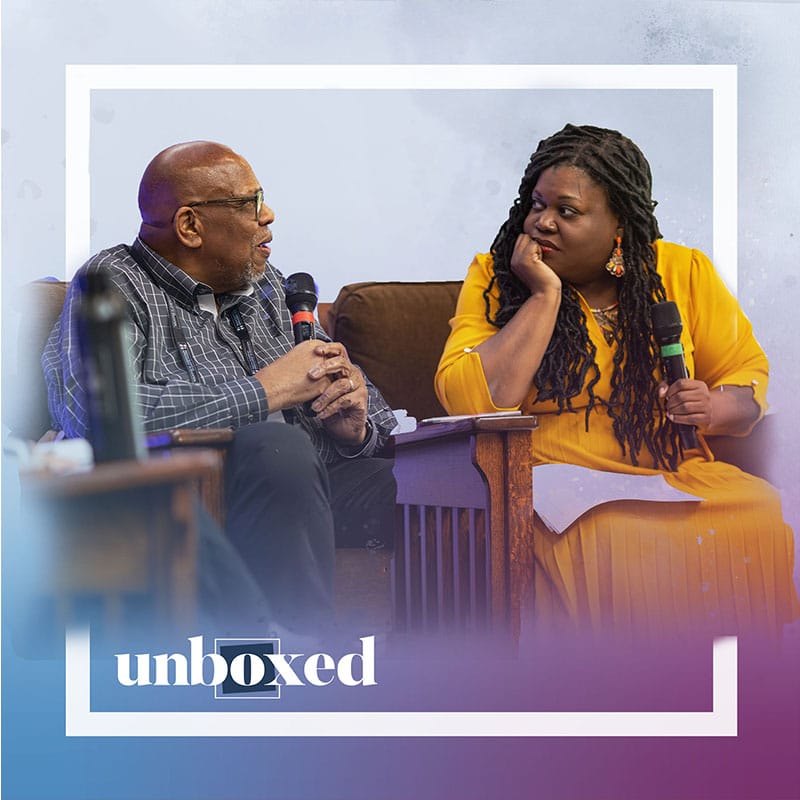 Two people sit and talk on a stage, each holding a microphone. The man on the left wears glasses and a plaid shirt. The woman on the right, Brandi Hinnant-Crawford, in a bright yellow dress, discusses promoting change. "Unboxed" is overlaid at the bottom.