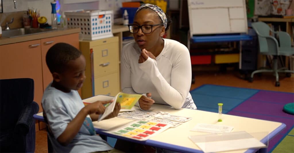 A woman in glasses sits at a table with an elementary student, watching as he points to a book during a student-led conference. Educational materials and a sand timer are on the table in a brightly lit classroom.