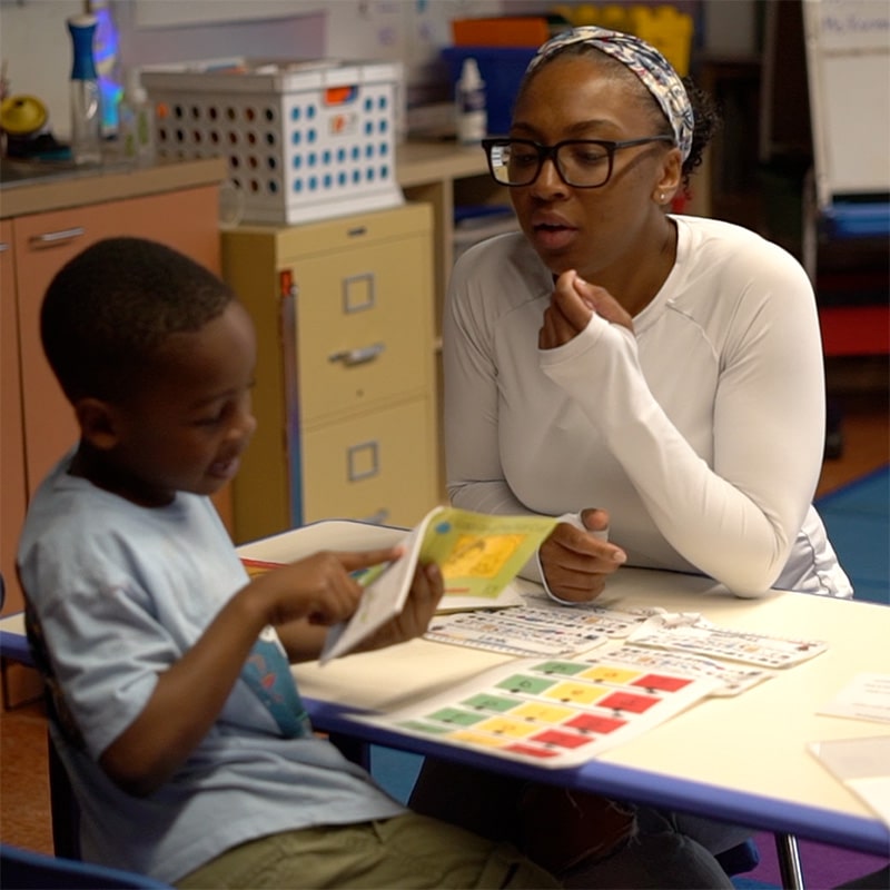A woman in glasses and a white top sits at a table with an elementary student, helping him read during a student-led conference. The boy points at the book, with educational materials spread out on the table in a classroom setting.