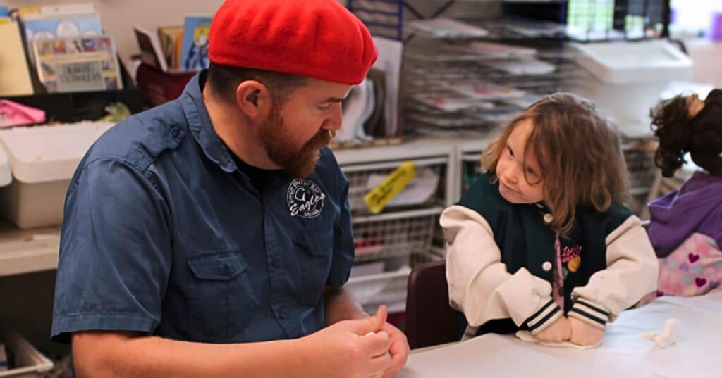A man in a red beret talks with a smiling young girl at a classroom table covered in white paper, surrounded by art supplies and shelves, fostering strong employee relationships through creative collaboration.