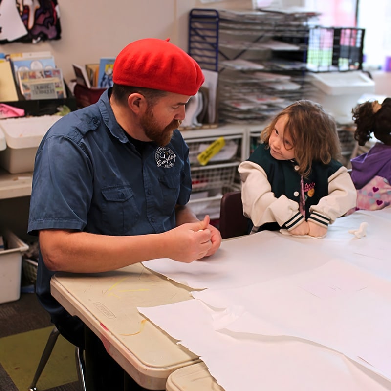 A man wearing a red beret and blue shirt sits at a table talking to a young girl in an art classroom. Surrounded by supplies, they smile, showing how creative spaces can strengthen employee relationships through shared experiences.