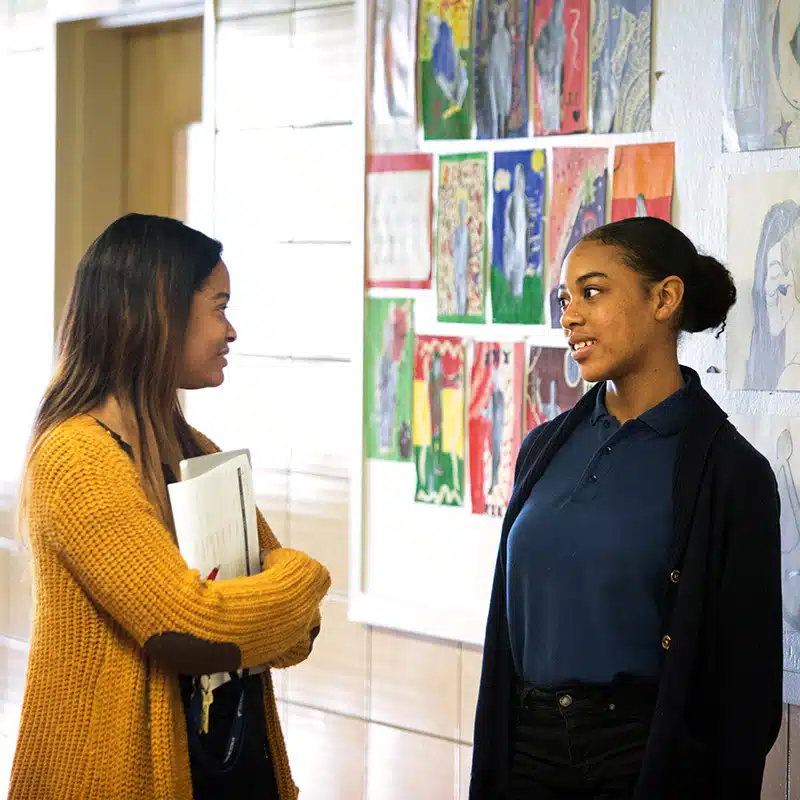 Two young women stand in a hallway talking about chronic absenteeism, one holding papers. Colorful artwork is displayed on the wall behind them. Both appear engaged in conversation.