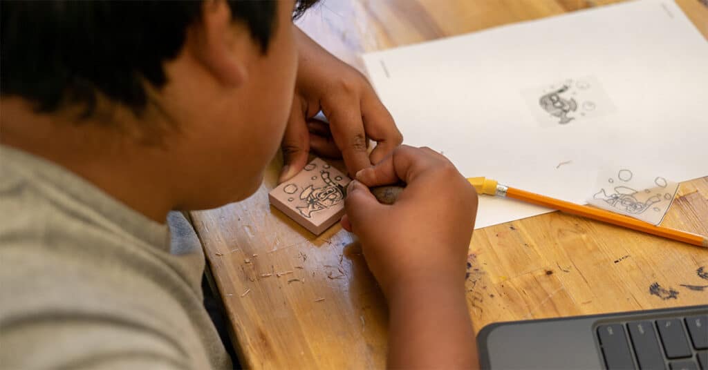 A person uses carving tools to engrave a design onto a small block, with a pencil, paper showing a sketch, and a laptop on the wooden table—blending creative soul with organized system in their workspace.