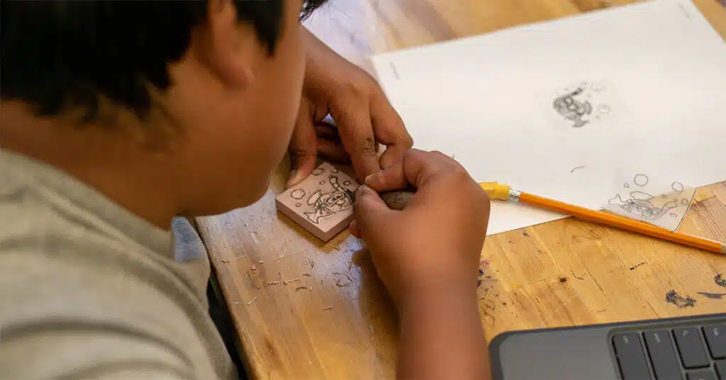 A person uses carving tools to engrave a design onto a small block, with a pencil, paper showing a sketch, and a laptop on the wooden table—blending creative soul with organized system in their workspace.
