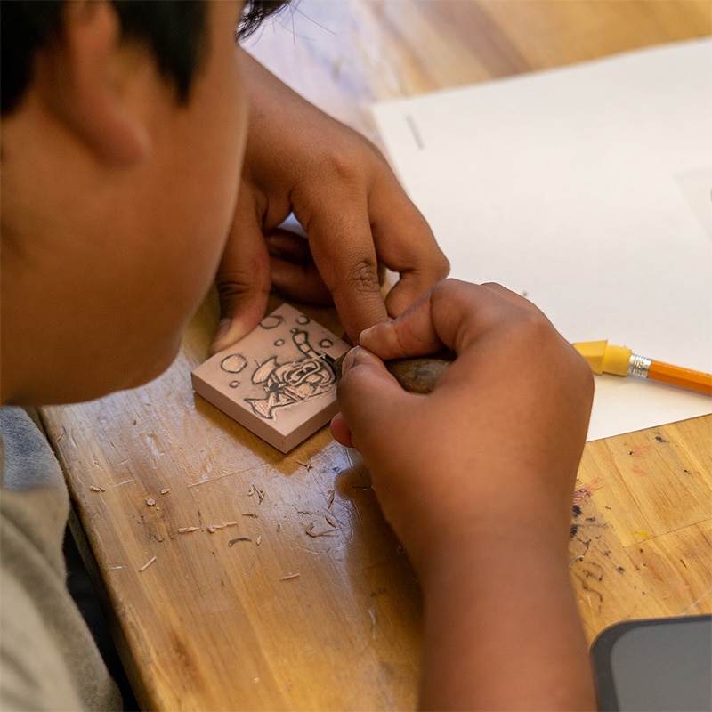 A person sits at a wooden table, carving a design into a small block with soulful focus, surrounded by wood shavings. A pencil and a piece of white paper are also on the table.
