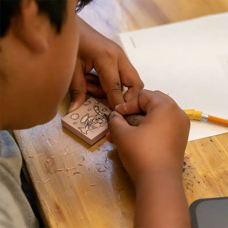 A person sits at a wooden table, carving a design into a small block with soulful focus, surrounded by wood shavings. A pencil and a piece of white paper are also on the table.