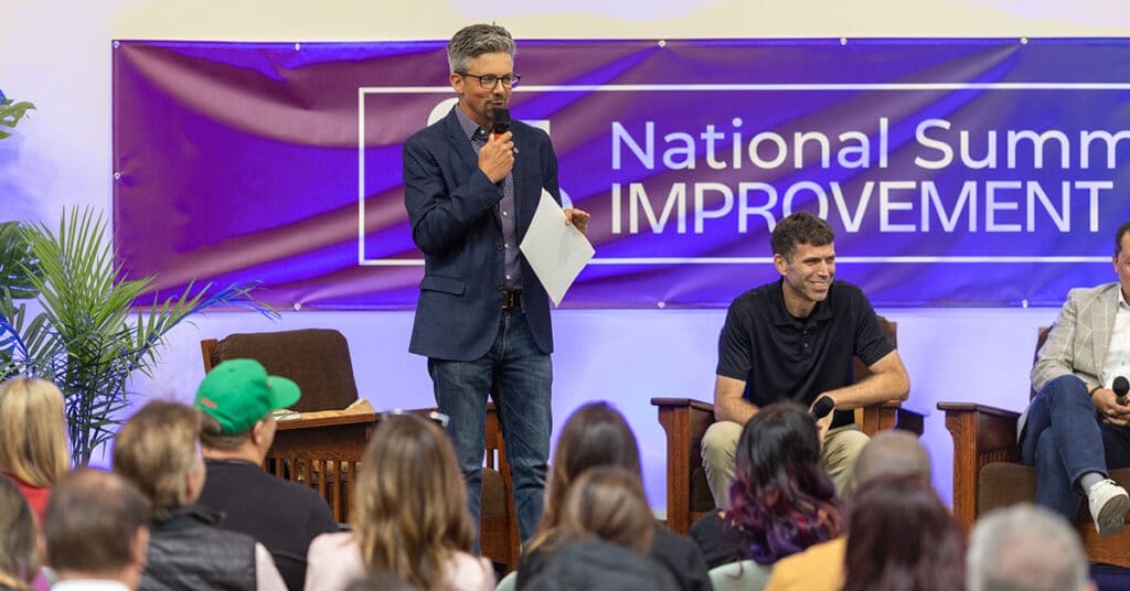 A man in a suit speaks into a microphone on stage at the National Summit for Improvement, featured in Unboxed Issue 30, while another man sits and smiles. An attentive audience watches them, with a blue banner visible in the background.