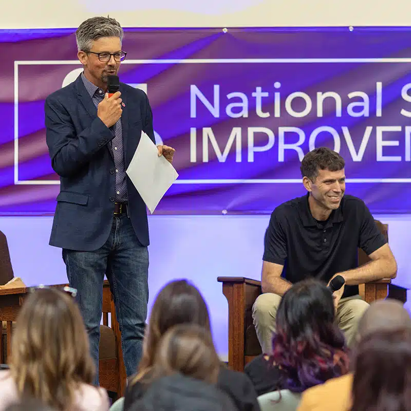 A man in a blue blazer speaks into a microphone while holding papers, standing next to a seated man in a black shirt, addressing an audience at the National S... event featured in Unboxed Issue 30.