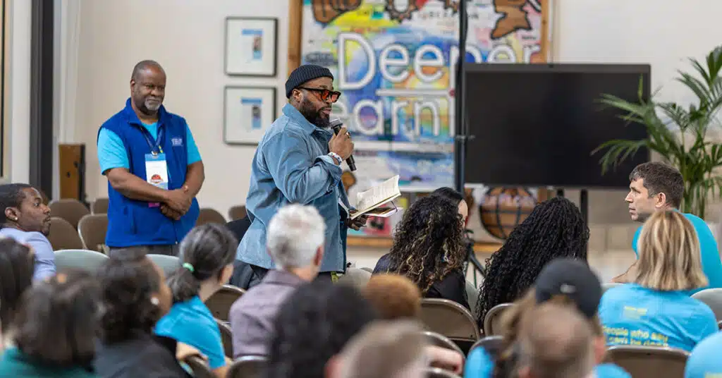 A man holding a microphone and notebook speaks to a seated audience, while another man in a blue vest stands nearby. Behind them, a colorful sign reads Deeper Learning—a system embraced by many attendees in matching blue shirts.