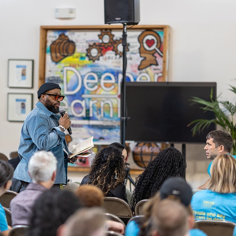 A man holding a microphone and a book speaks to an audience seated indoors. Behind him is a colorful sign reading Deeper. The atmosphere feels like a casual seminar or cypher, with several people in matching blue shirts.