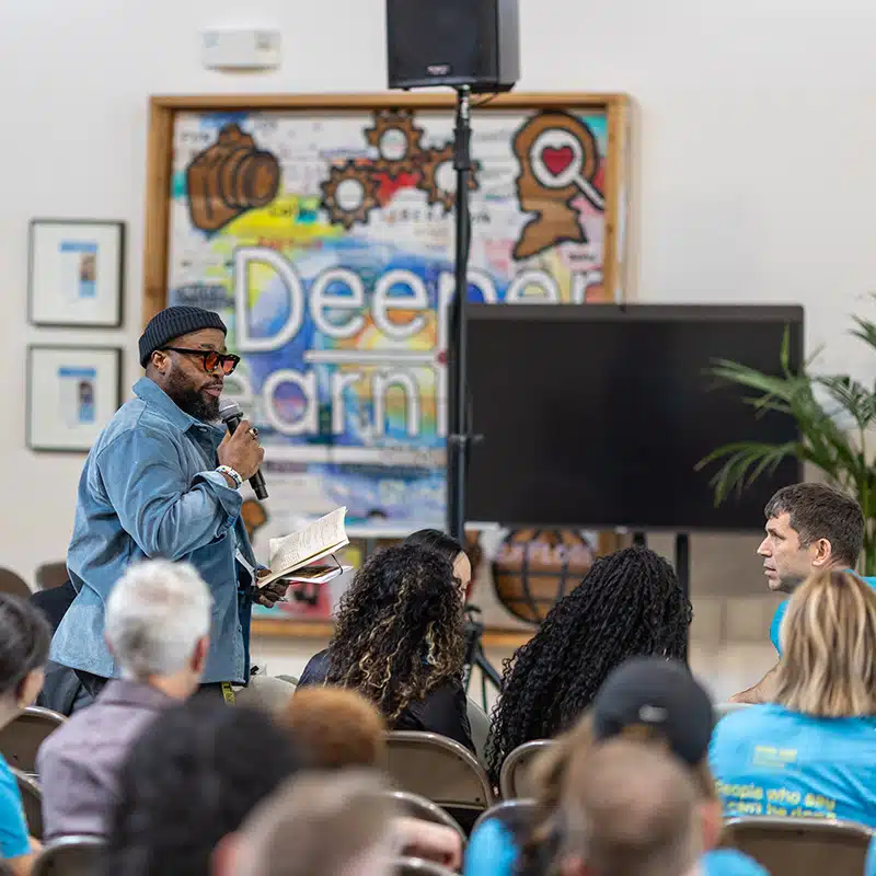 A man holding a microphone and a book speaks to an audience seated indoors. Behind him is a colorful sign reading Deeper. The atmosphere feels like a casual seminar or cypher, with several people in matching blue shirts.