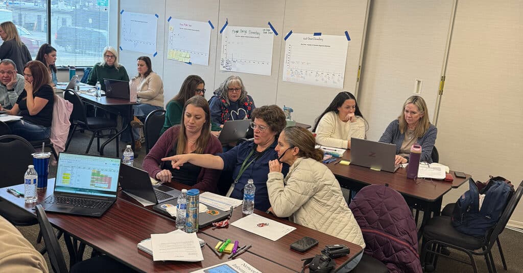 A group of adults sits at tables in a classroom, working together on laptops. Some people are collaborating and pointing at screens, discussing strategic communication, while charts and notes on reducing absenteeism are posted on the wall behind them.