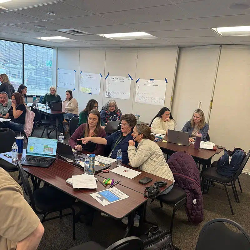 People sit at tables in a conference room, collaborating and working on laptops. Papers, water bottles, and jackets are on the tables. Large sheets of paper with notes about reducing absenteeism are taped to the walls in the background.