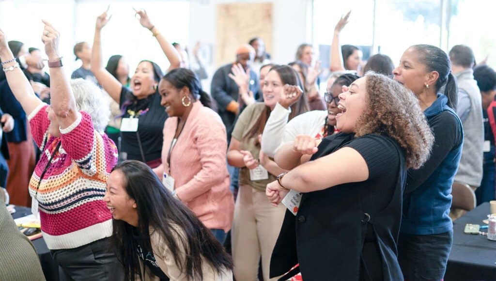 A diverse group of people stand together indoors, smiling, cheering, and raising their arms enthusiastically in a lively, celebratory atmosphere that highlights team collaboration and a shared commitment to continuous improvement.