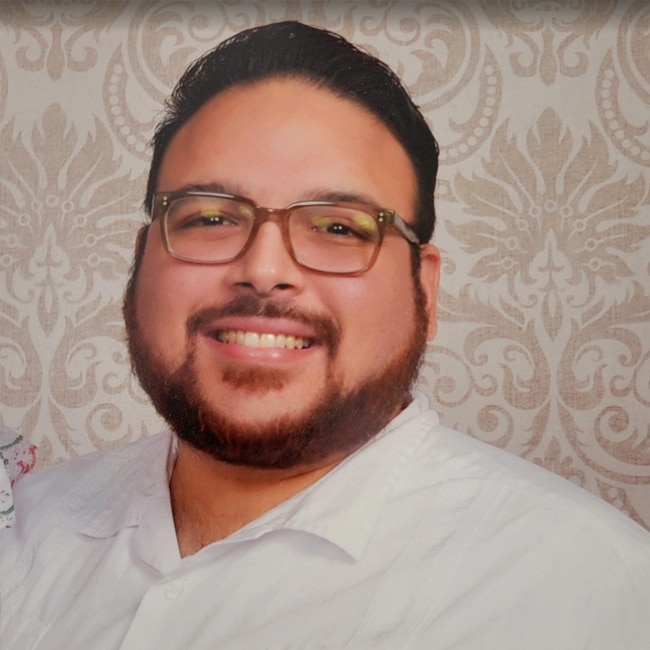 Smiling man with short dark hair, beard, and glasses wearing a white shirt, posing in front of a light patterned wallpaper background. This is Ernesto Orozco Jr.
